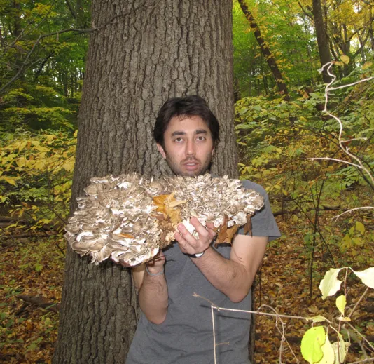 Photo of David Horvitz in the forest holding up a huge pile of mushrooms.