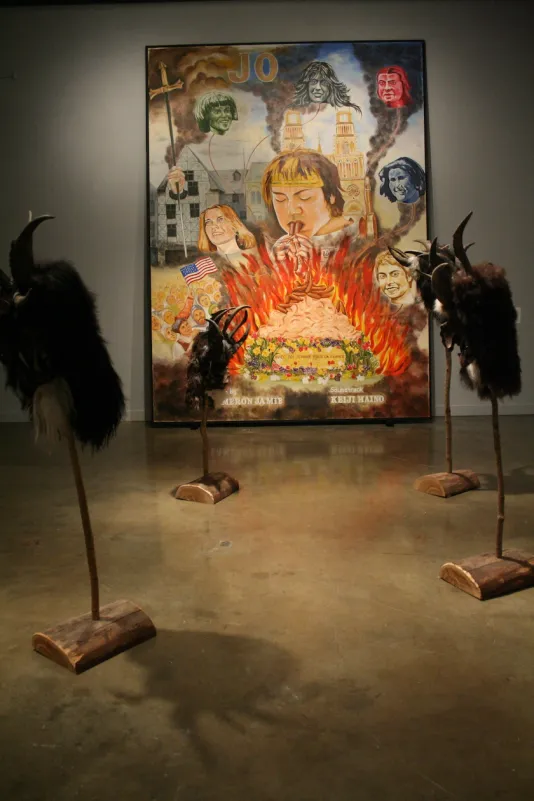 Large colorful painting of heads and fire facing 5 bestial horned masks on wood stakes with sketches on the right wall.