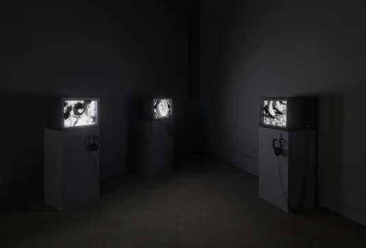 Three televisions are on display in a gallery space on display podiums. They are all playing videos of black and white shapes.