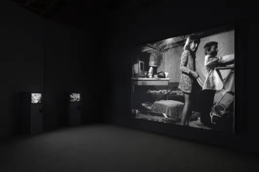 Black and white photo of video projection and three chairs. Projection features two children in a cluttered room with a desk and computer. 