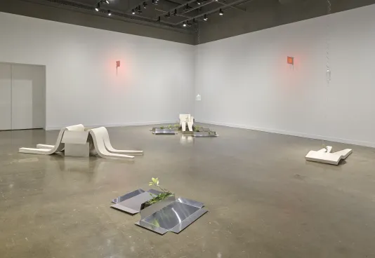 A gallery at the MIT List Visual Arts Center features sculptures consisting of white plaster slumps, aluminum sheets, weeds, and red glowing signs on the walls.