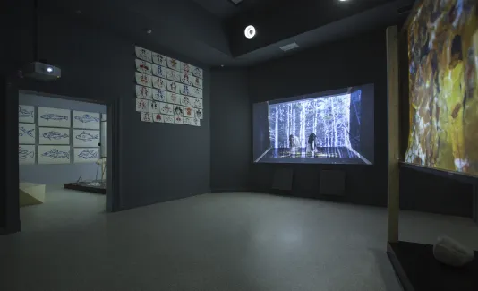 Dark room with projectors on two walls and prints go beed on the left wall.