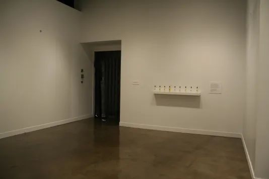 Installation view of a white gallery with a small shelf that has plastic perfume bottles on it.