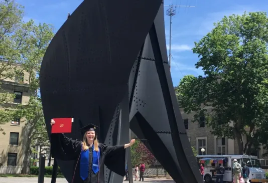 A girl in a graduation gown holding up a red diploma stands in front of a large, black steel sculpture