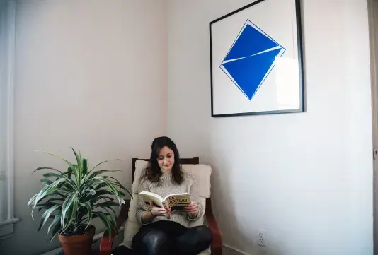 A woman reads in a chair in the corner of a room with houseplant to her right and a blue silkscreen print to her left.