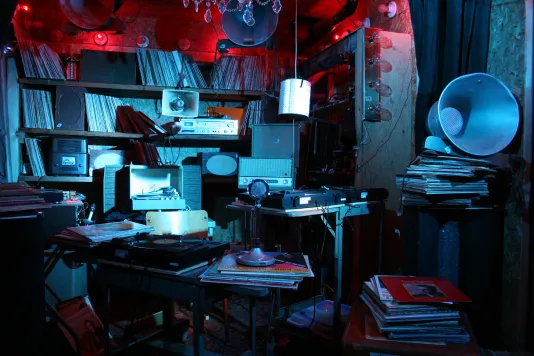 Installation view of  blue lit room cluttered with items such as records and decorations.