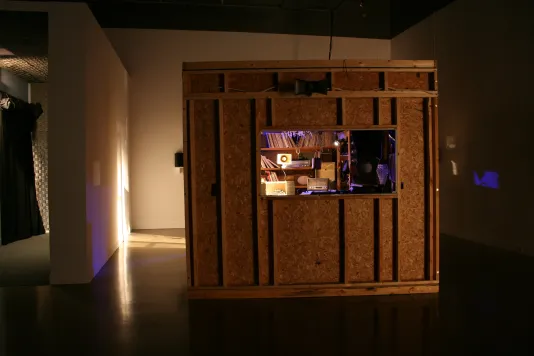 Installation view of a crate in a gallery with a hole inside peaking into a small lit up room filled with records.