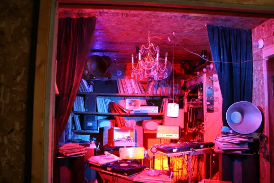 Installation view of a room inside a crate lit with red lights featuring cluttered albums and decorations.