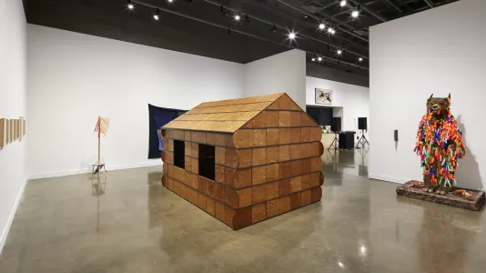 Installation view featuring a wooden cabin sculpture in the foreground