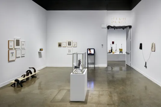 Installation view of a gallery featuring framed prints on the walls and small objects inside a glass container in the center of the room.