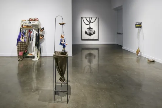 Installation view featuring a sculpture of a puppet with a clothing rack in the background.