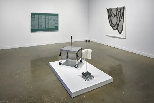 Installation view featuring a metal card on a podium in the center, a green print with white text on the wall, and a tapestry with chain like graphic on the right wall.