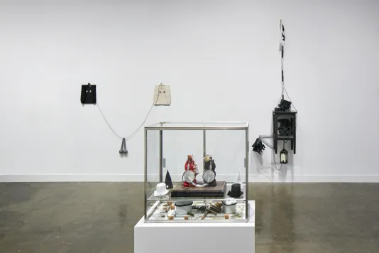 Installation view featuring a glass box with small objects inside and sculptures hanging on the wall behind.