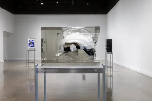 Installation view featuring a white helmet encased in a rectangular resin mold on a metal stand and two similar style sculptures in the background. 