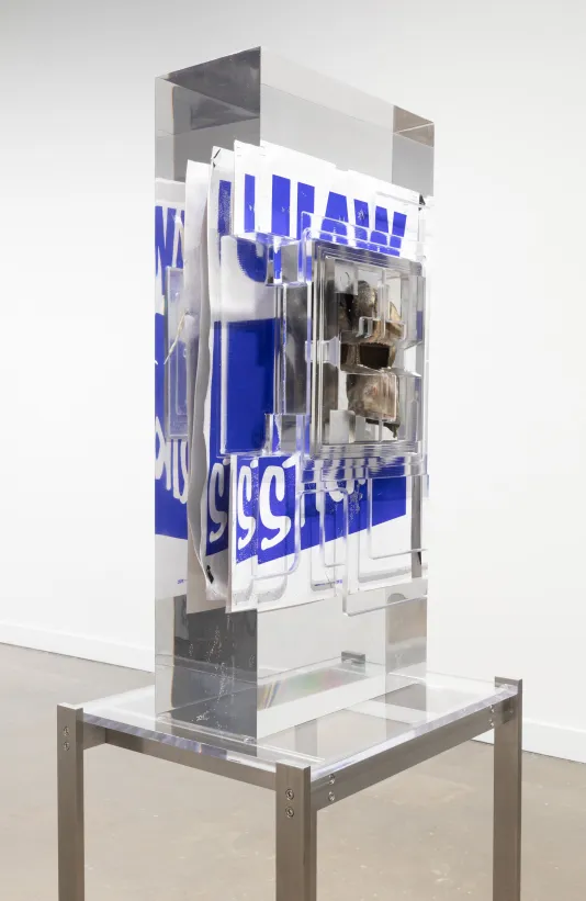 Installation view of a work glove in front of a blue and white poster that reads “UAW” on strike encased in a block of resin on a metal stand.