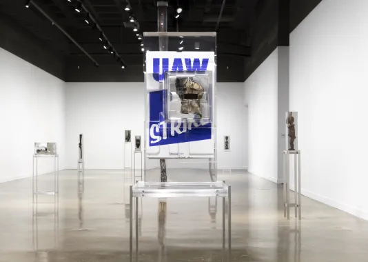 Installation view featuring a work glove in front of a blue and white poster that reads “UAW” on strike encased in a block of resin on a metal stand.