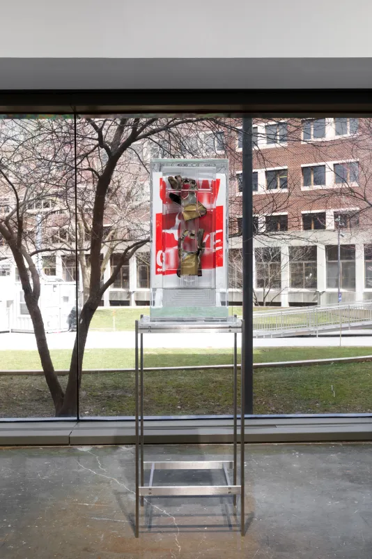 Installation view of a pair of work gloves and a red poster encased in a rectangular block of resin on a metal stand with a large window overlooking a courtyard in the background. 