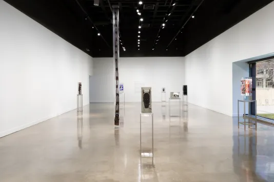 : Exhibition view featuring around 8 sculptures of varying sizes with objects inside of blocks of clear resin spread throughout the gallery. Each sculpture is on a tall metal stand.