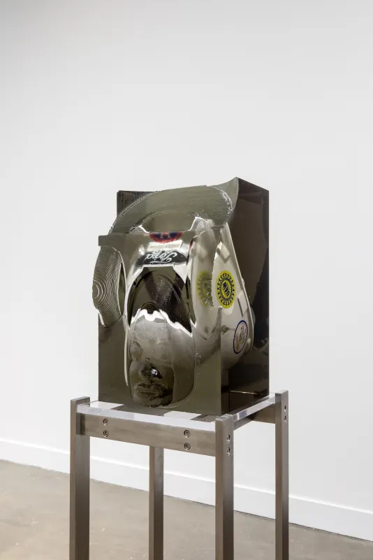 Installation view featuring a white Ford helmet with stickers on the side encased in a block of resin on a metal stand. 