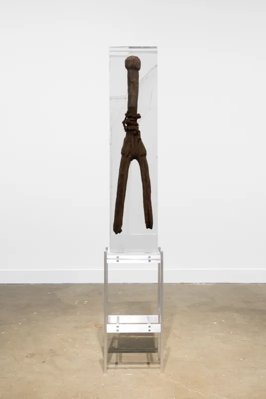 Installation view of a tall wooden sculpture with a round top and two long legs encased in a block of resin on a metal stand.