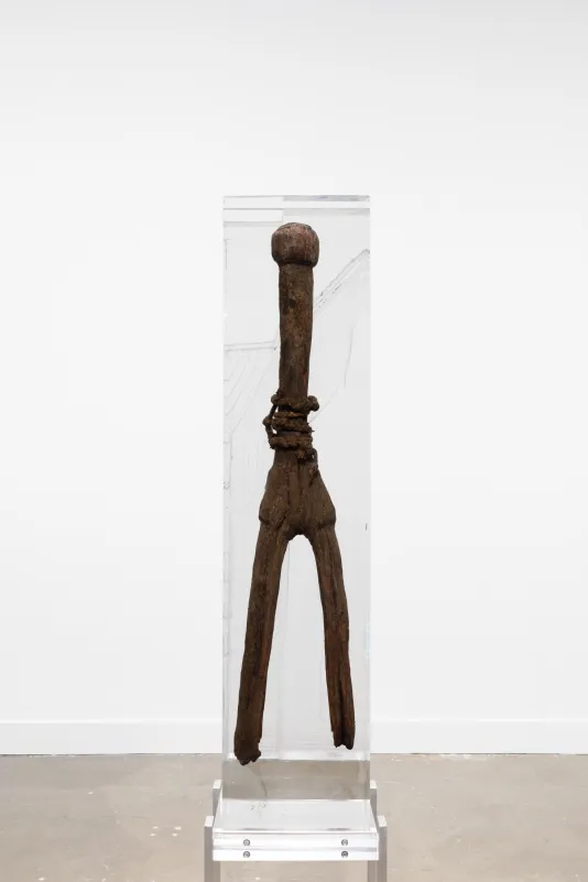 Installation view of a tall wooden sculpture with a round top and two long legs in a block of resin on a metal stand.