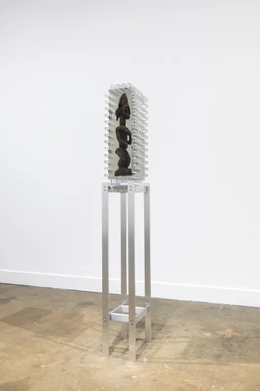 Installation view of an African wooden sculpture of a figure encased in a block of resin with jagged edges on a tall metal stand. 
