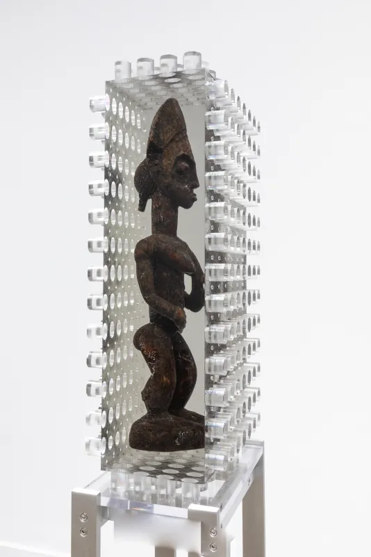 : Installation view of an African wooden sculpture of a figure encased in a block of resin with jagged edges on four of the six sides. 