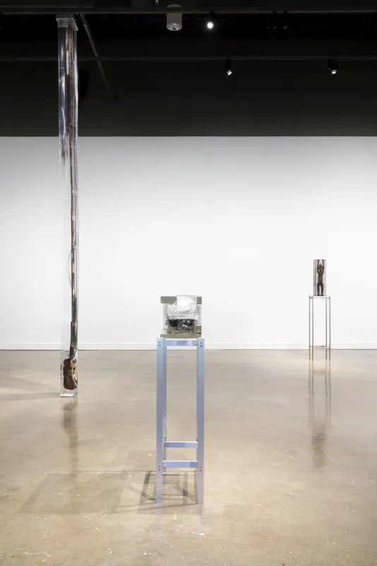Exhibition view featuring two sculptures with objects inside of blocks of clear resin on a tall metal stand on the right and a floor to ceiling sculpture inside resin on the left.