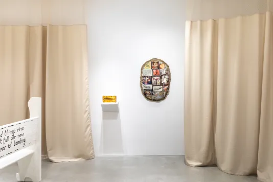 Installation view featuring a white church pew with black text, a gelatin sculpture containing a dried fish and an oval shaped textile sculpture hanging on the wall, surrounded by warm tan anti-microbial medical curtains. 