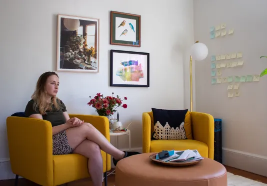 A woman sits in an office sitting area on one of two yellow chairs with three framed artworks behind her.
