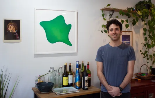 A man poses with his hands together smiling in front of a piece of artwork that has a green abstract shape. There is a bar cart and plant in the background.