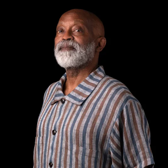 A brown skinned man with a full white beard stands with hands behind his back in front of black backdrop. He wears a striped shirt