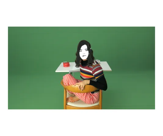 Video still of a person from behind sitting at a desk turned towards the camera with a cartoon looking face cropped onto her and a green screen background.