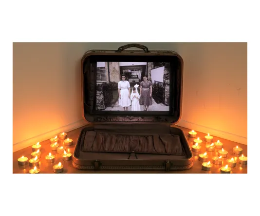 Video still of a suitcase propped open with a photograph from a first communion inside taking up the width of the suitcase. Tealight candles light the room on the ground to the left and right of the case.