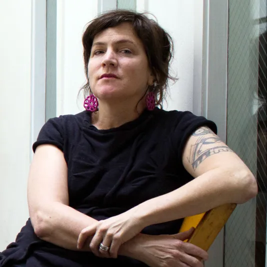 Lexa Walsh, seated in a wooden chair, wears a black t-shirt, bright pink earrings. She has brown hair pulled back and fair skin