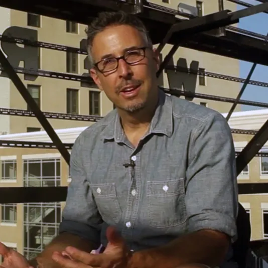 Rubin sitting outside wearing a blue button-up with sleeves rolled up and dark frame glasses. He has short hair and facial hair