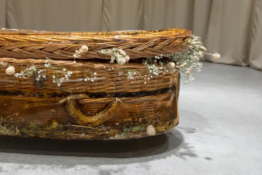 Detailed view of a wicker casket encased in resinous gelatin with objects including syringes, pills, and beans on its side, and dried flowers surround the opening between its lid and base.