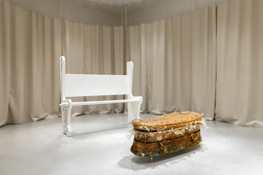 Sculpture of a willow wicker child size casket cast in gelatin sits on the floor in front of a white painted wooden church pew with beige hospital curtains lining the gallery walls.