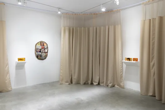Beige hospital curtains line the gallery wall with gelatin sculptures on a white shelf and an oval shaped textile sculpture hanging on the wall.