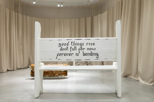 A painted white church pew from behind with the quote “good things rise don’t fall for now forever is bending” written on the back in black in a gallery surrounded in beige hospital curtains.