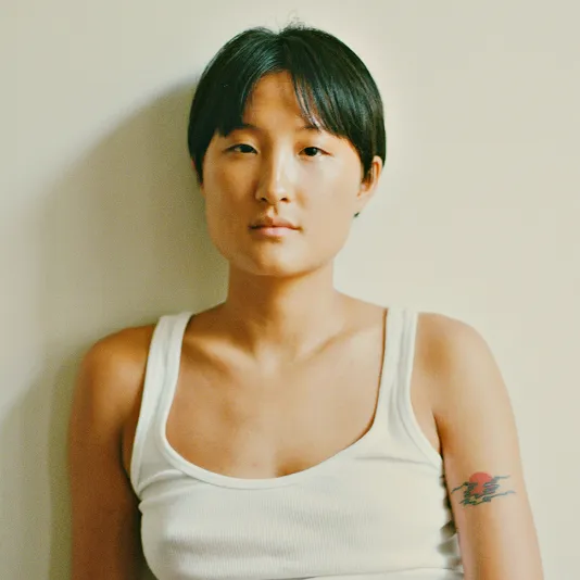 Artist TJ Shin with short, dark hair sits on a chair with legs crossed, in front of a blank wall. Shin wears a tank top and blue jeans
