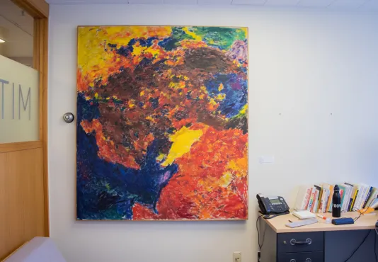 A large square painting hangs on an office wall with a desk to the right and a wooden wall to the left.