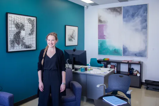 A young woman wearing all black stands in her office with a teal wall on the left, two framed artworks on the left wall and one large artwork on the back wall. 