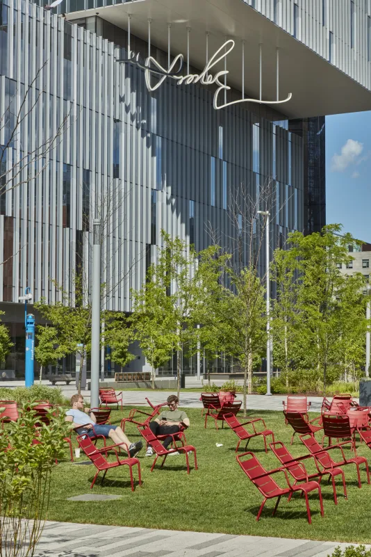 Lawn with red steel chairs in the foreground with a building in the back and newly installed LED sculpture of a hand drawn signature hanging down from the buildings overhand.