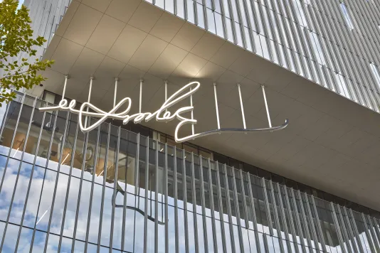 White LED sculpture of a signature being lit up hanging from a glass building. 