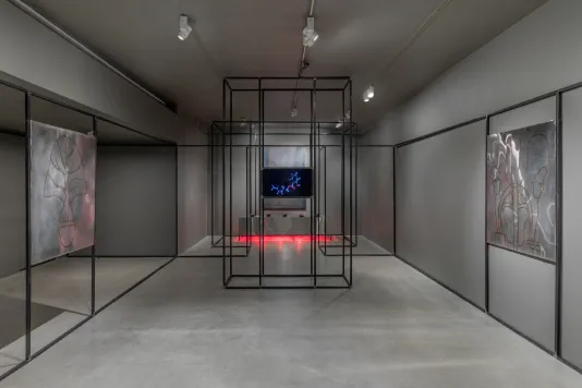 Gallery view with grey walls and cement flooring featuring welded steel drawings on the left and right, a monitor in the center, a steel platform at rear glows red from heat lamps underneath, and a black steel architecture snakes along the periphery of the space.