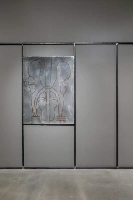 : A welded steel drawing sits in between two vertical steel bars on a gallery wall surrounded by other steel bars that form rectangular shapes on the wall. 