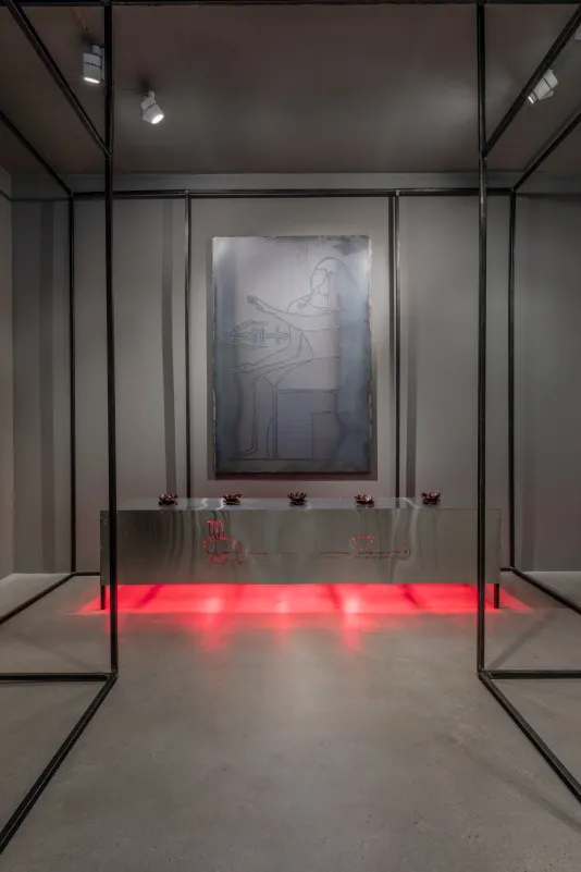 Black steel architecture frames a metal table against the rear gallery wall with red heat lamps lighting it from underneath and five small bukhoor sculptures on top with a large, welded steel drawing of a Pharaoh hanging above the table. 