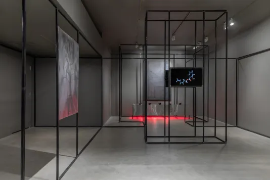 Gallery view with grey walls and cement flooring featuring welded steel drawings on the left and right, a monitor in the center, a steel platform at rear glows red from heat lamps underneath, and a black steel architecture snakes along the periphery of the space.  