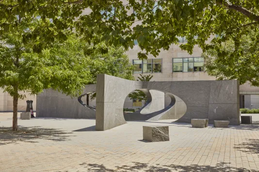 Granite open air sculpture featuring a rectangular shape with ovals cut out in the middle surrounded by trees peaking through the top edge of the photo. 